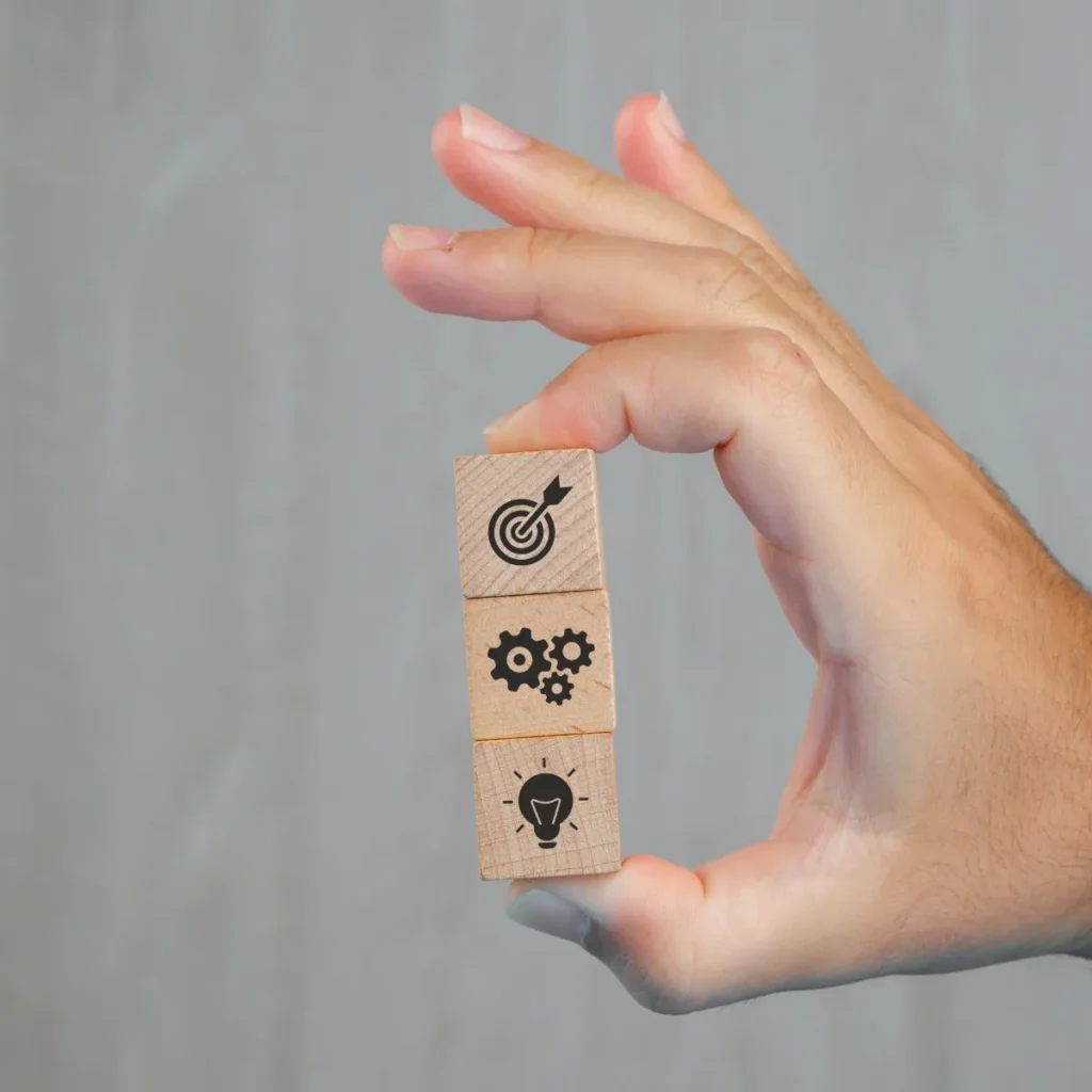 Hand holding three wooden blocks with icons of target, gears, and lightbulb - Frost Flare